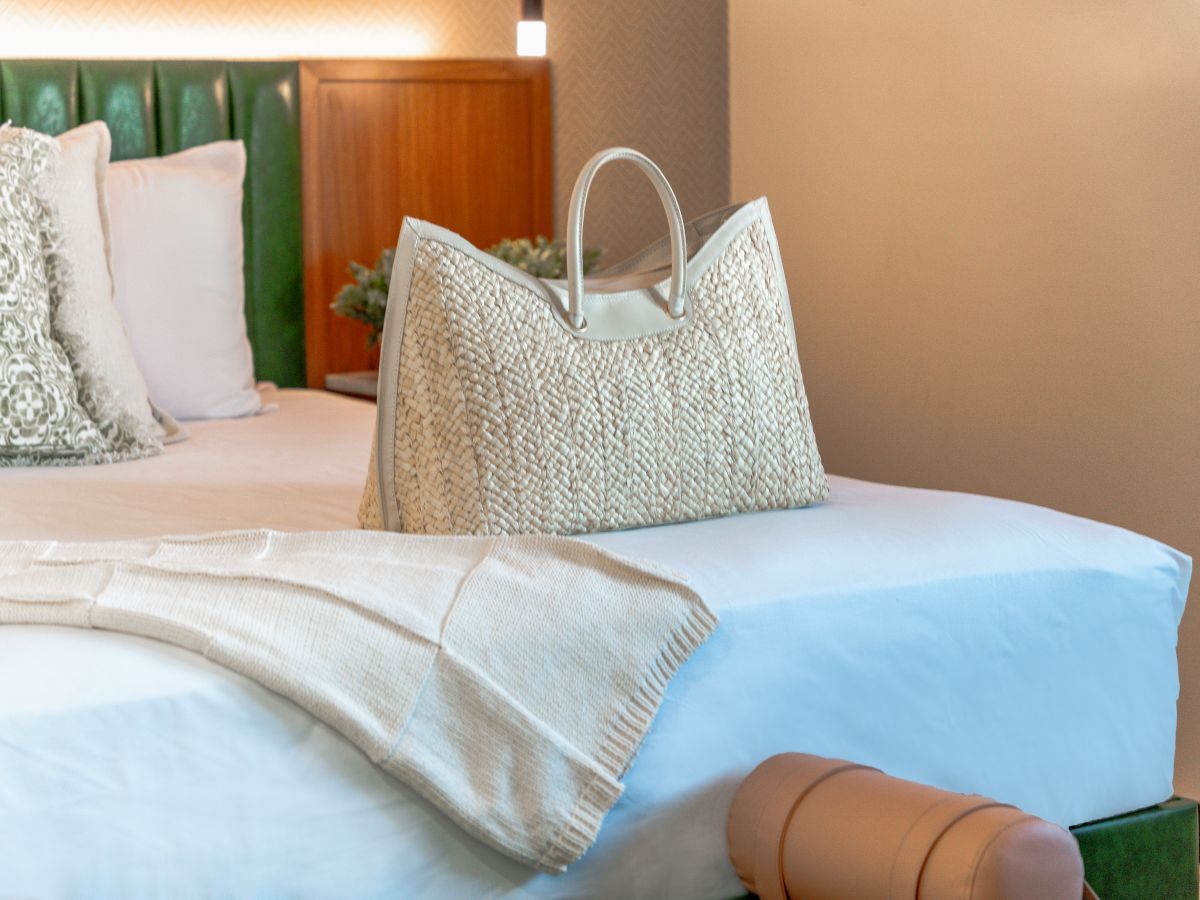 A close-up of a bed with white bedding, a green headboard, and a woven bag on top.