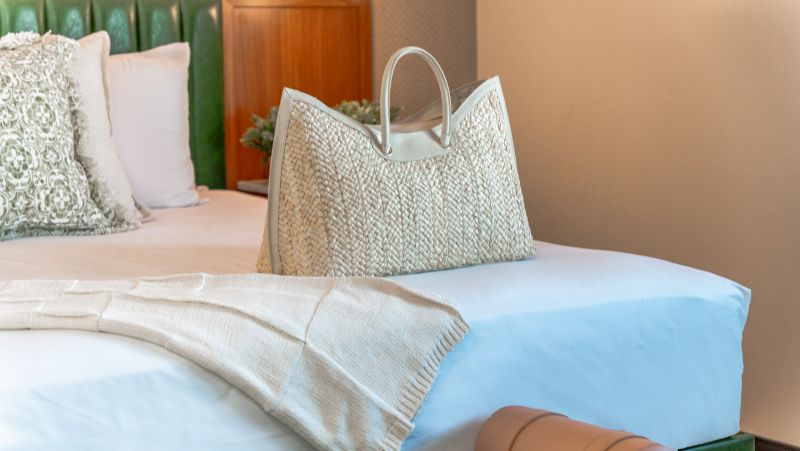 A close-up of a bed with white linens, a green headboard, and a woven handbag.