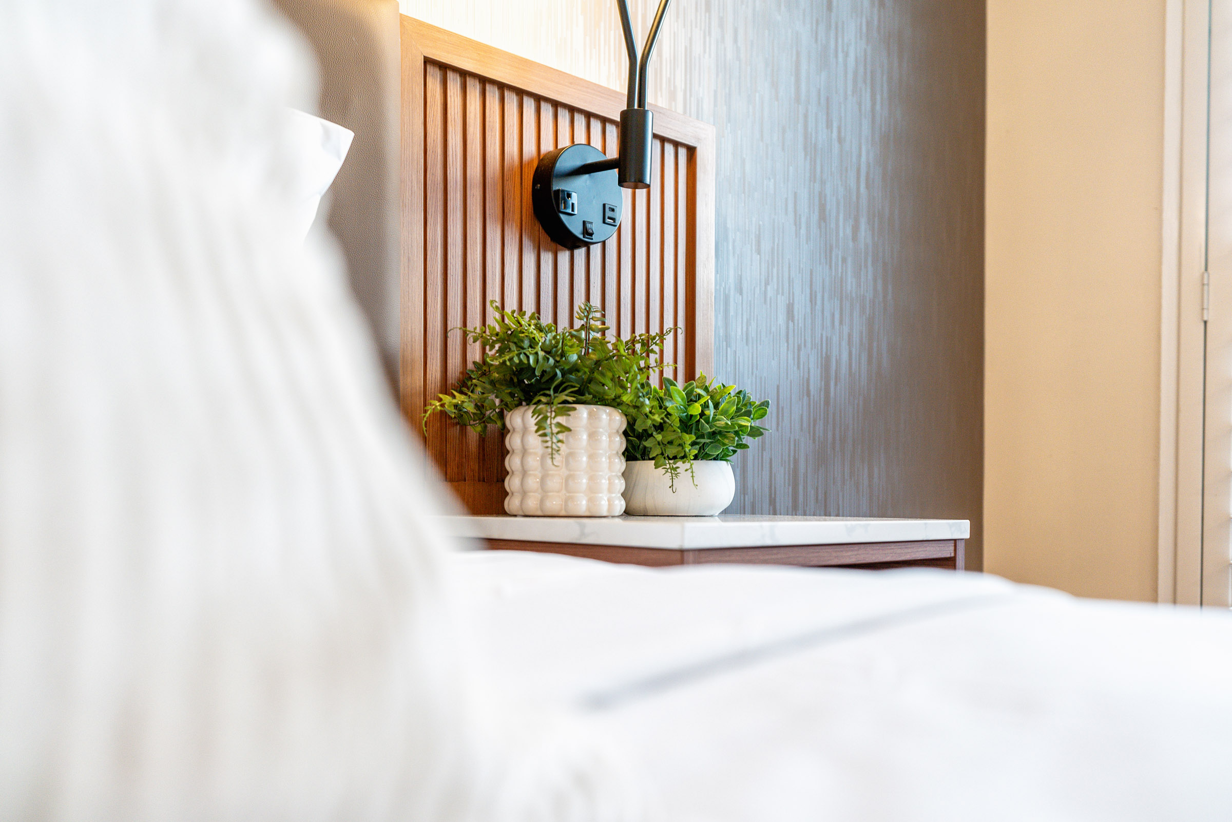 A close-up of a bed with a wooden headboard, white bedding, and a marble nightstand with two potted plants and a black lamp.
