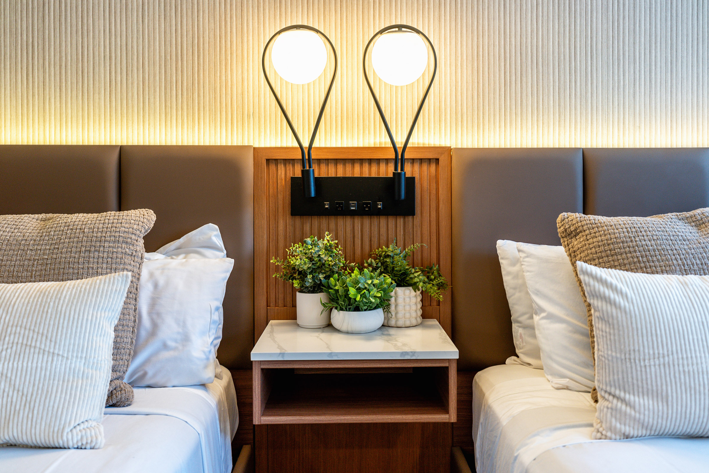 A close-up view of a hotel room with two beds, a wooden nightstand with a marble top, and potted plants.