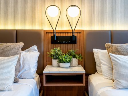 A close-up view of a hotel room with twin beds, a wooden nightstand with a marble top and potted plants, and reading lamps.