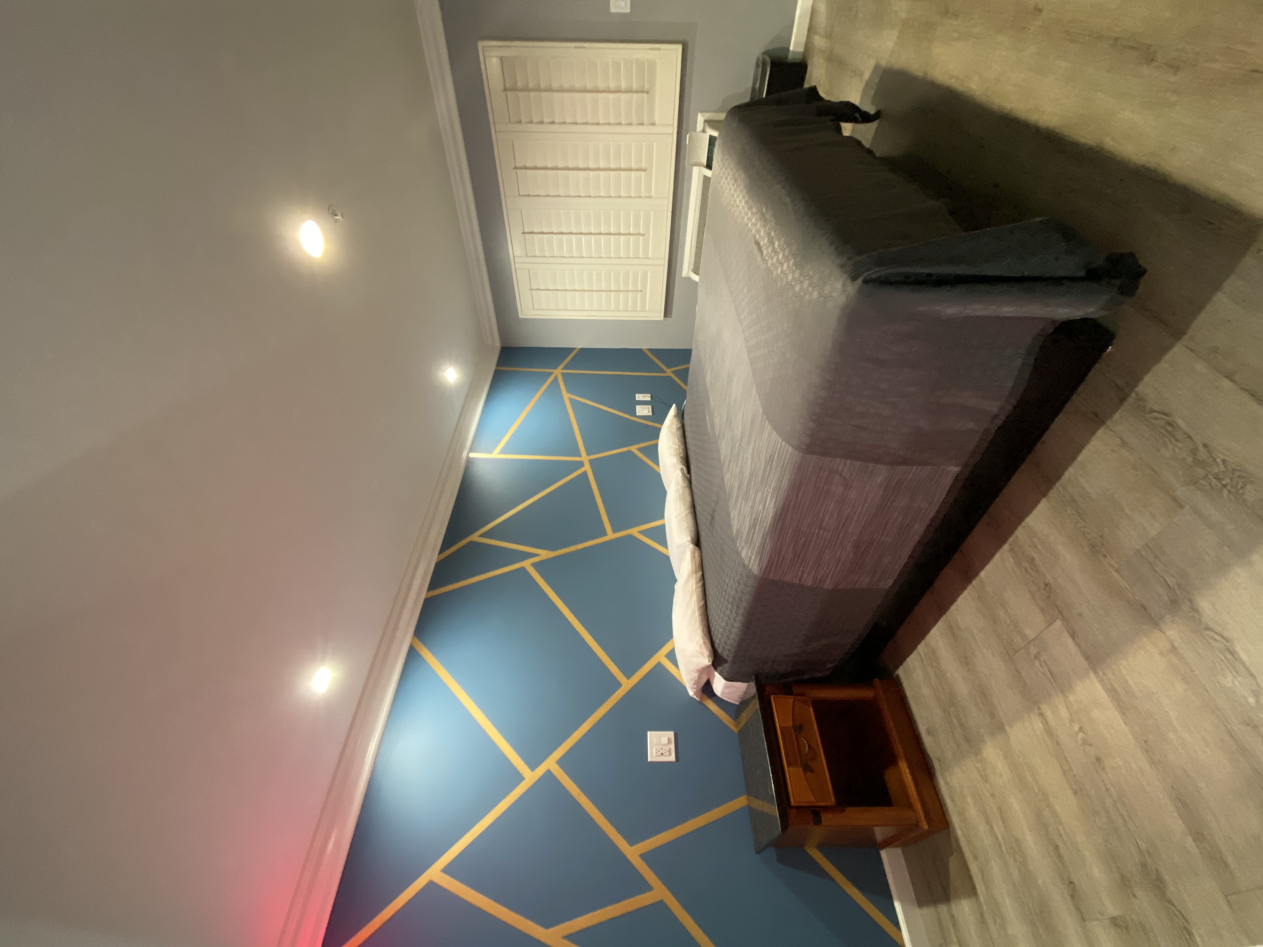 A guest room with a blue floor with yellow lines, a gray bed, and a white door.