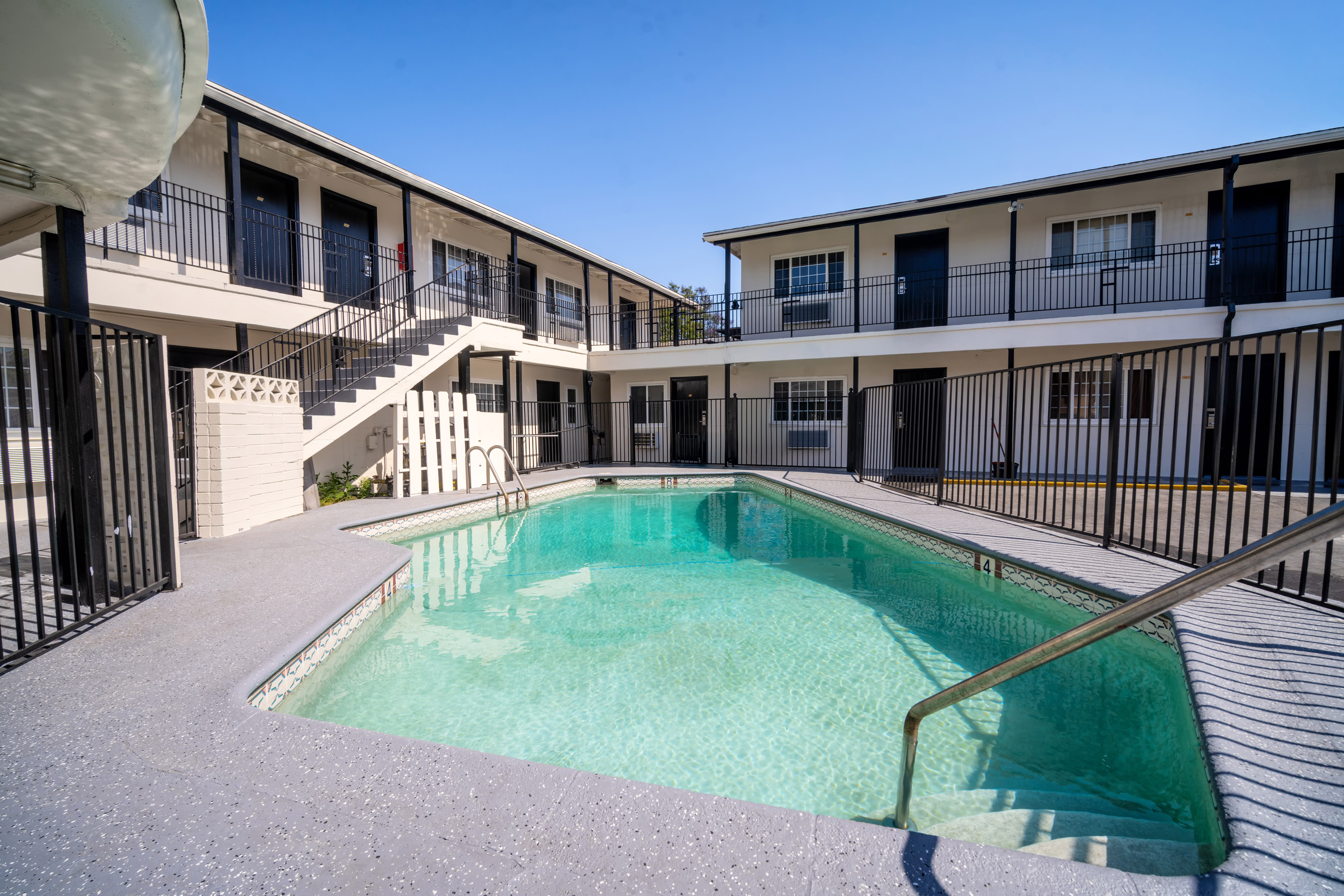 A rectangular pool with a metal handrail and concrete deck surrounded by a building with balconies and stairs.